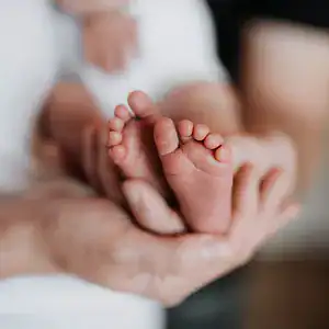 Close-up of an adult gently cradling a newborn's tiny feet in both hands. The scene conveys warmth, tenderness, and protection. Background is softly blurred.