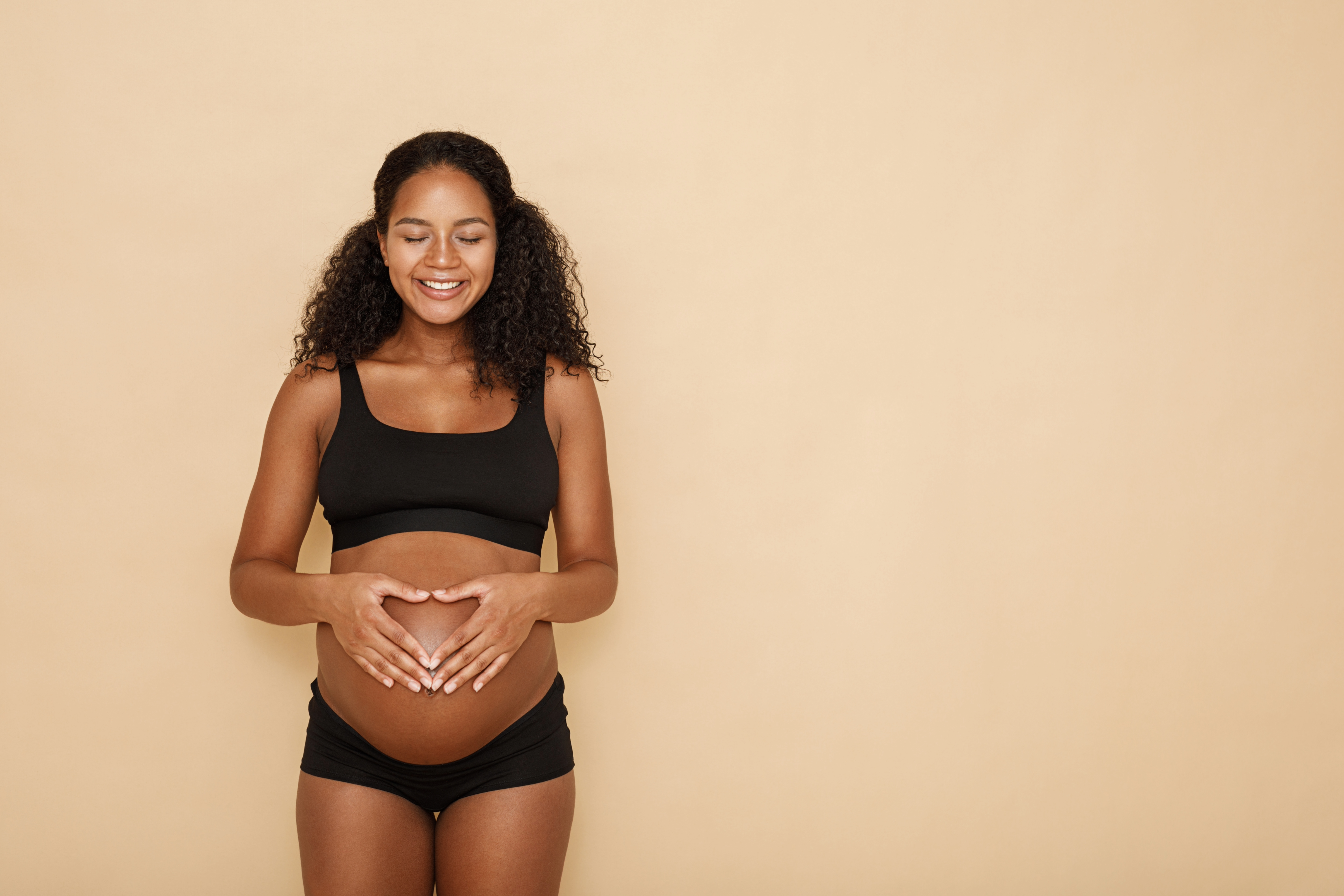 Pregnant woman with curly hair in black underwear, eyes closed, smiling, forms heart on belly with hands against beige background, expressing joy.
