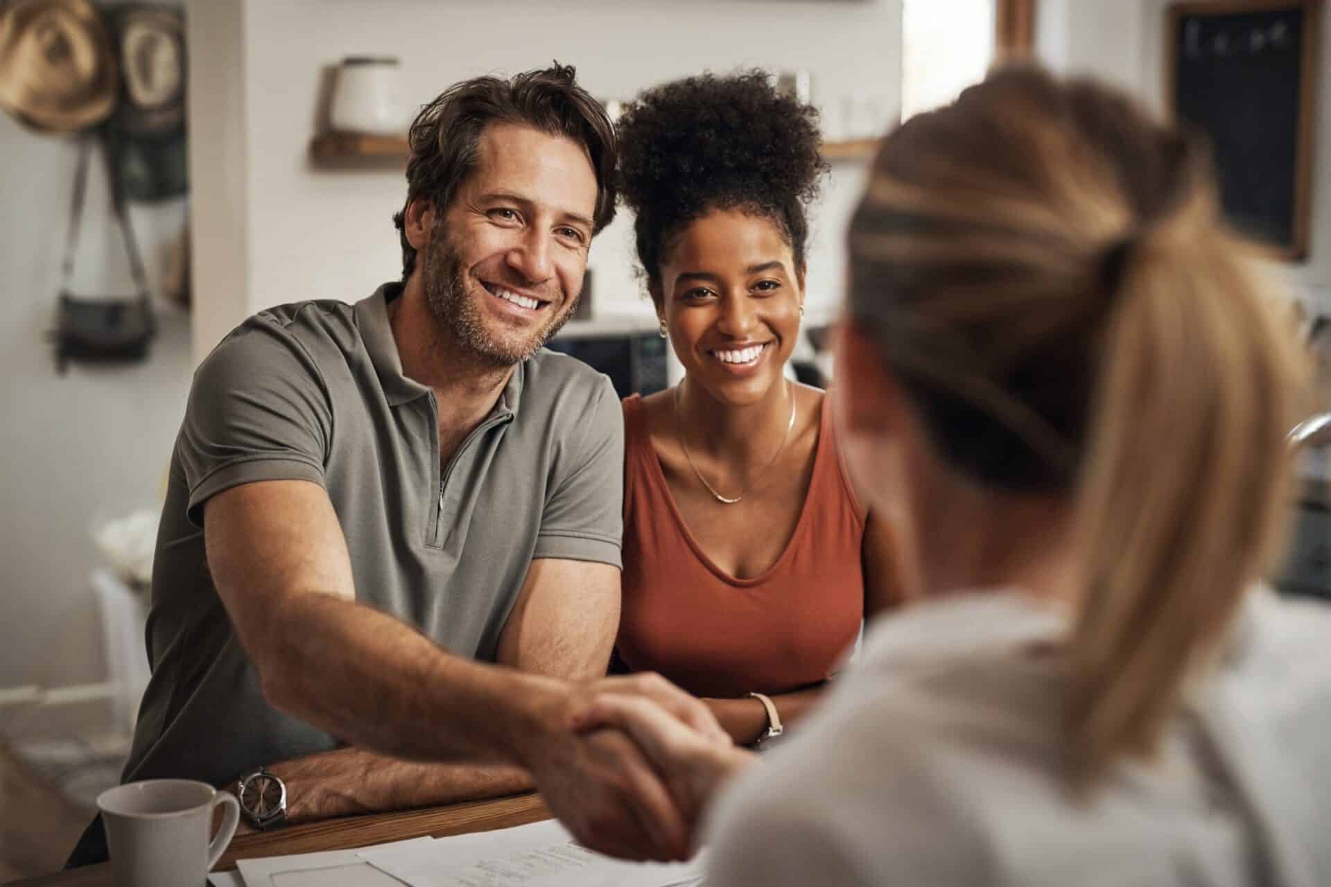 A smiling couple sits at a table, shaking hands with a person whose back is to the camera, conveying a friendly, welcoming atmosphere.