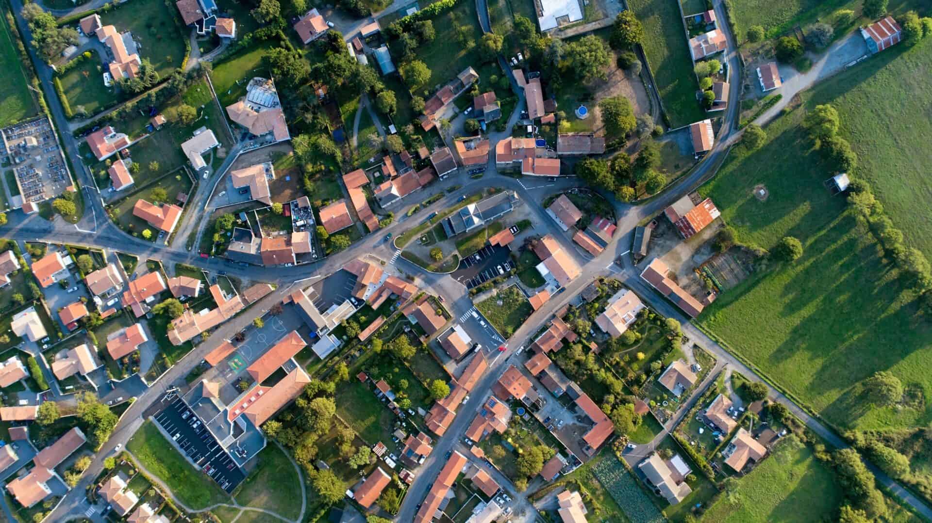 Aerial view of a village with red-roofed houses, winding roads, and lush green fields, conveying a peaceful, rural setting under soft sunlight.