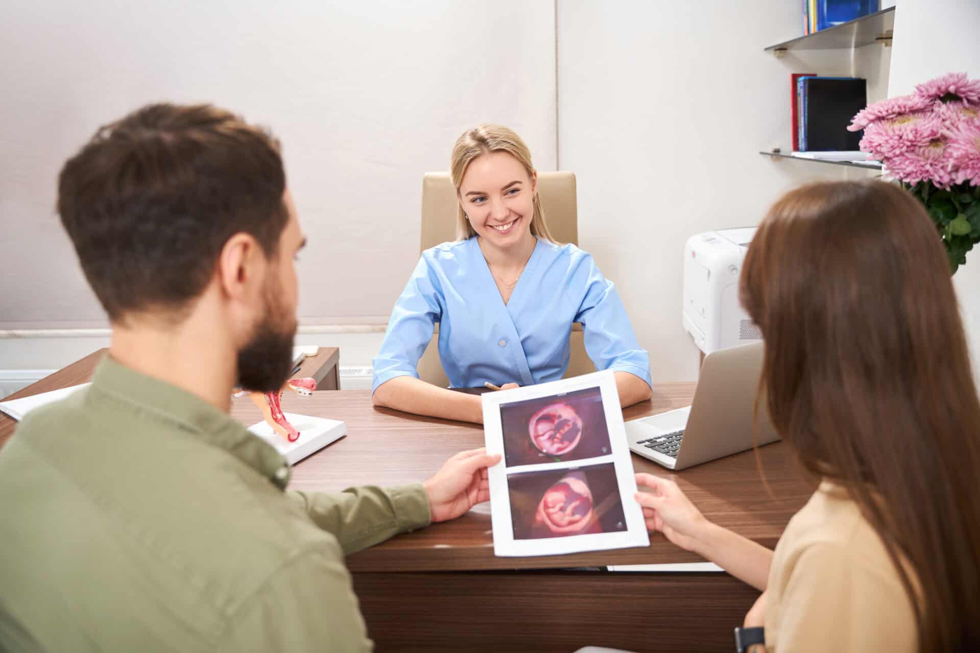A smiling doctor in light blue scrubs sits at a desk, engaging with a couple holding ultrasound images. The atmosphere is warm and supportive. Best Surrogacy Agencies