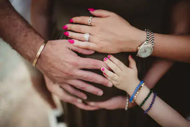 Hands with jewelry gently rest on a pregnant belly, symbolizing family unity and anticipation. The scene conveys warmth and expectation.