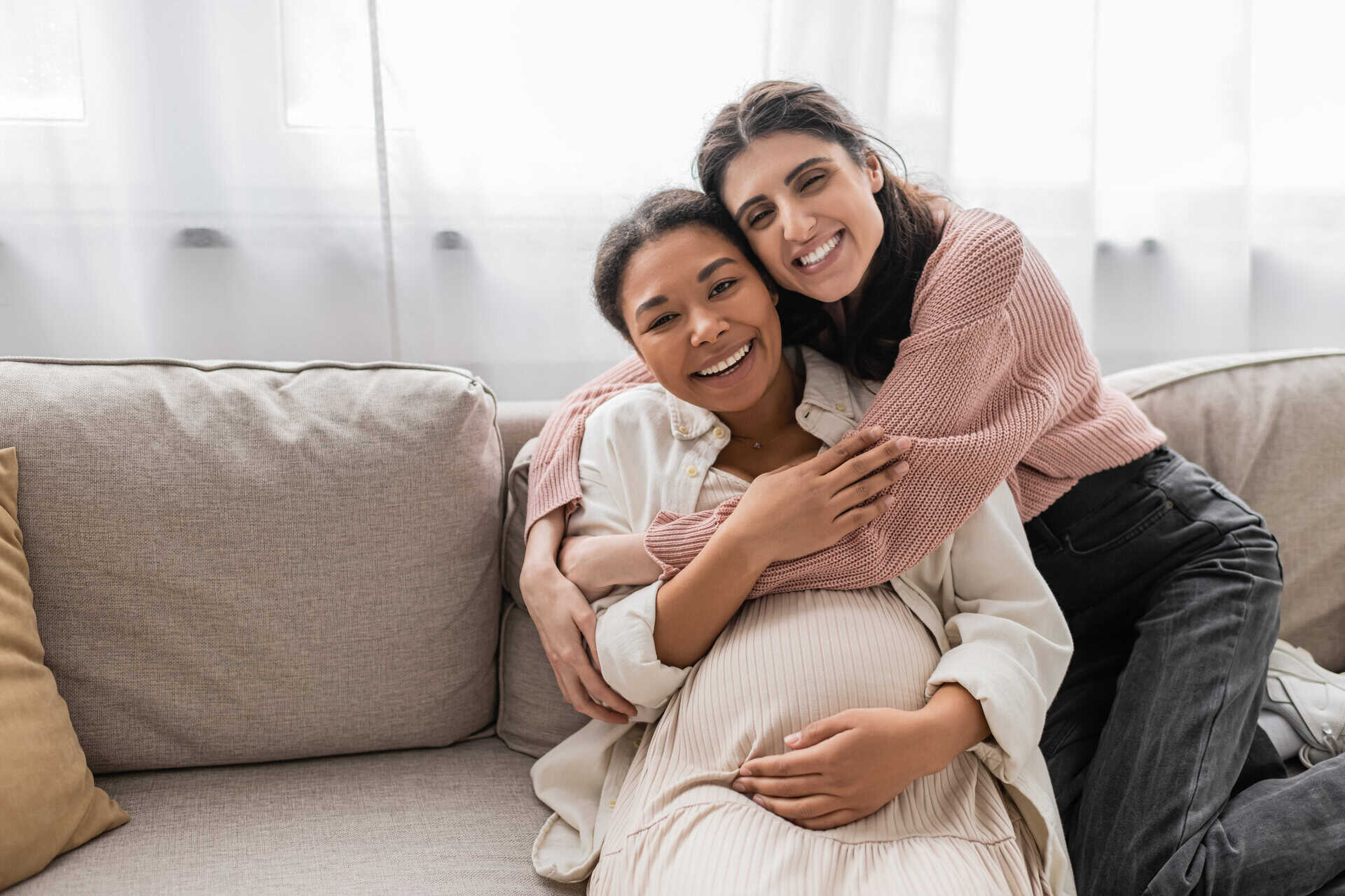 Two women smiling warmly on a sofa, with one hugging the other from behind. The seated woman cradles her pregnant belly, conveying joy and affection.