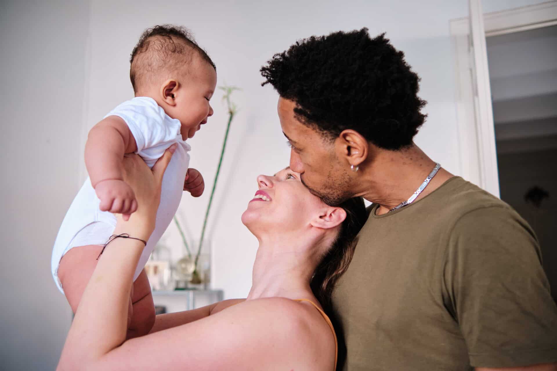 A couple joyfully holds up a laughing baby in a white onesie. The father tenderly kisses the mother's forehead, radiating warmth and family love. How to Find a Surrogate Mother
